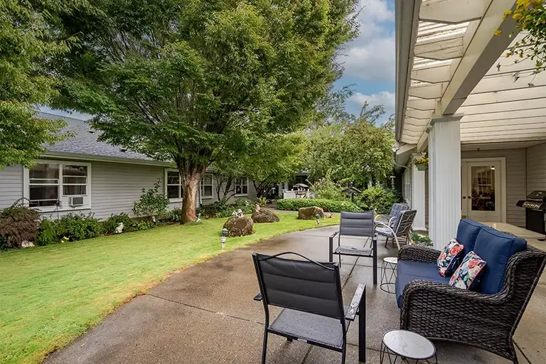 A patio with outdoor seating overlooks a grassy yard with trees and shrubs, adjacent to a light gray Trustwell Senior Living house under a partly cloudy sky. A patio with outdoor seating overlooks a grassy yard with trees and shrubs, adjacent to a light gray Trustwell Senior Living house under a partly cloudy sky.