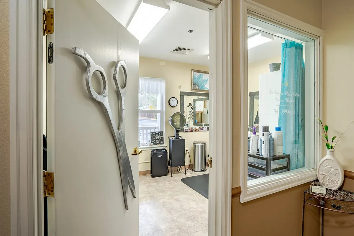 Open door with large decorative scissors leads into the Trustwell Senior Living hair salon, featuring mirrors, chairs, hair products, and a window that fills the space with natural light.