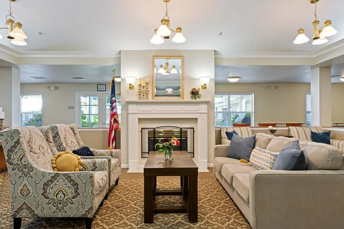 A well-lit living room at Trustwell Senior Living features patterned armchairs, a beige sofa, a wooden coffee table, a cozy fireplace, and an American flag proudly displayed in the background.