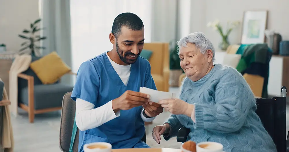 A caregiver in blue scrubs sits beside an elderly woman in a wheelchair, both smiling and looking at a piece of paper together at a table with drinks and snacks.