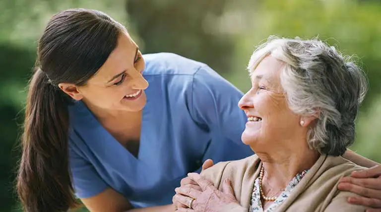 A caregiver in blue scrubs smiles at an older woman, who is sitting and smiling back, outdoors with blurred greenery in the background at Trustwell Senior Living. A caregiver in blue scrubs smiles at an older woman, who is sitting and smiling back, outdoors with blurred greenery in the background at Trustwell Senior Living.
