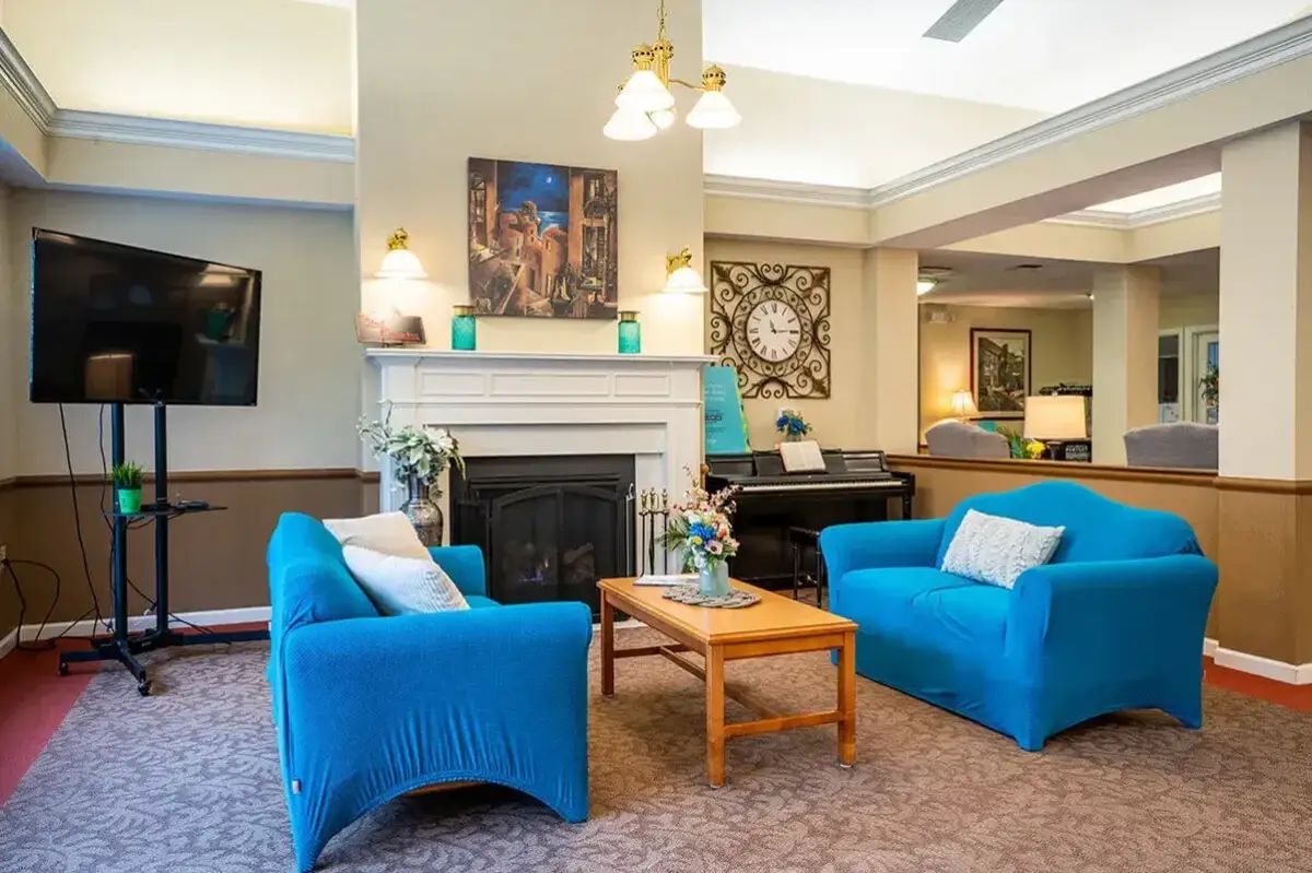 A cozy living room at Trustwell Senior Living features two blue armchairs, a wooden coffee table, a fireplace, a wall clock, a TV on a stand, and a piano, all beautifully decorated with lamps, flowers, and framed art.