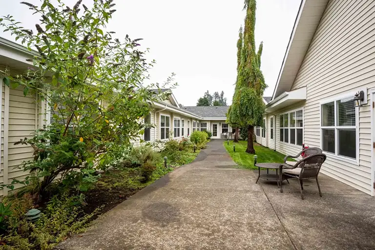 A concrete walkway runs between two beige Trustwell Senior Living buildings with windows, lined with bushes and trees, and features a seating area with two chairs and a small table. A concrete walkway runs between two beige Trustwell Senior Living buildings with windows, lined with bushes and trees, and features a seating area with two chairs and a small table.