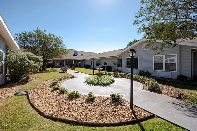 A well-maintained courtyard at Trustwell Senior Living features a circular walkway, a central flower bed, inviting benches, and surrounding single-story buildings under a clear blue sky. A well-maintained courtyard at Trustwell Senior Living features a circular walkway, a central flower bed, inviting benches, and surrounding single-story buildings under a clear blue sky.