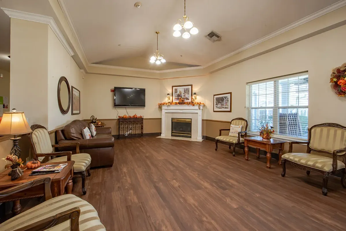 A spacious, well-lit living room at Trustwell Senior Living features wooden floors, a brown leather sofa, armchairs, a cozy fireplace, wall-mounted TV, and warm autumn decorations on tables and the mantle.