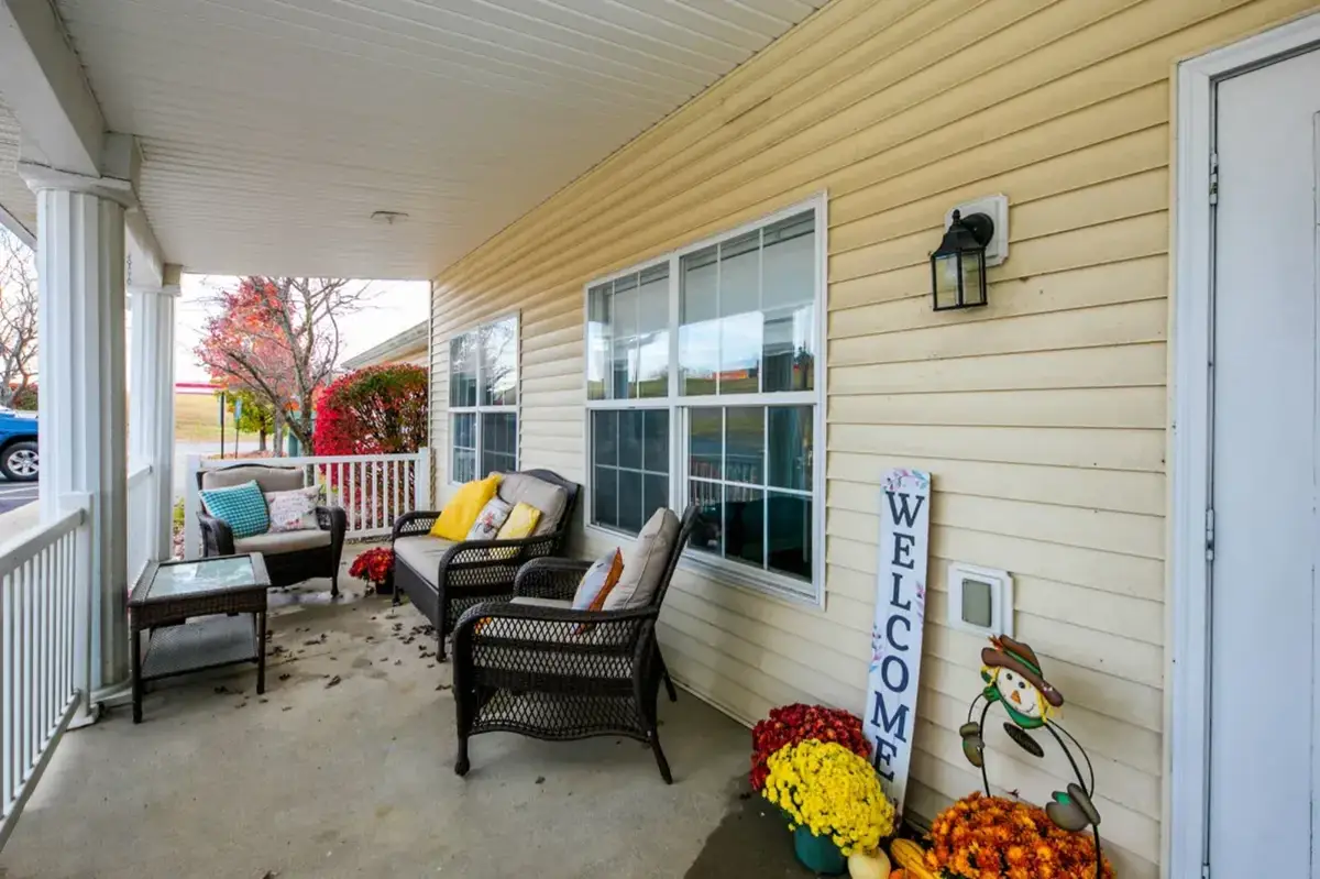 A front porch at Trustwell Senior Living features wicker furniture, colorful cushions, a glass-top table, potted flowers, a "Welcome" sign, and a small scarecrow decoration near the door.