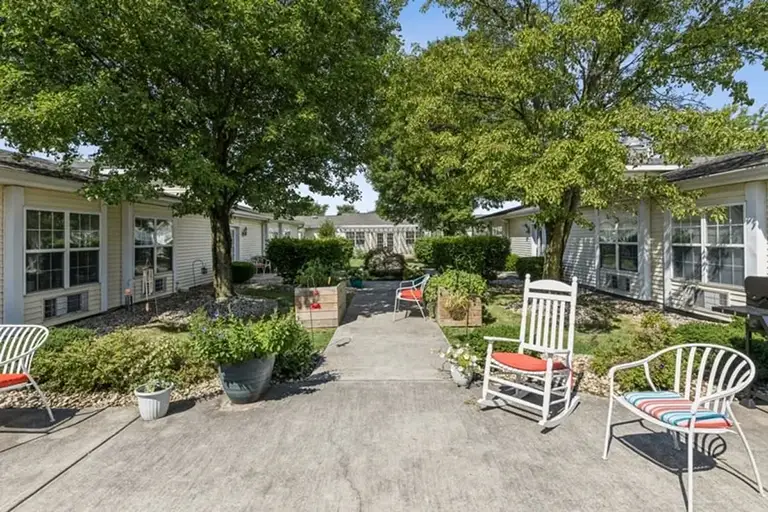 Trustwell Senior Living features an inviting outdoor courtyard with a concrete walkway, garden beds, and several chairs, including rocking chairs with red cushions, all set amid single-story buildings and shady trees. Trustwell Senior Living features an inviting outdoor courtyard with a concrete walkway, garden beds, and several chairs, including rocking chairs with red cushions, all set amid single-story buildings and shady trees.