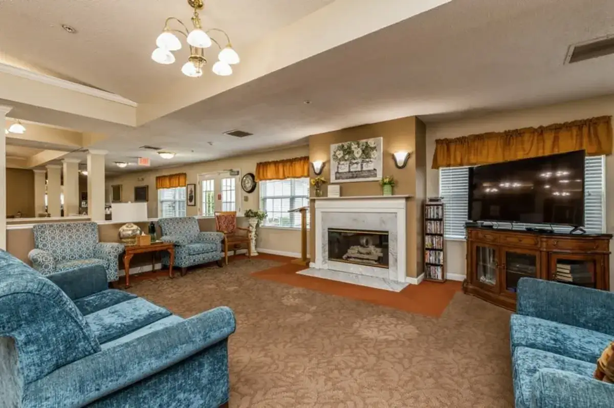 A cozy living room at Trustwell Senior Living features blue sofas, armchairs, a fireplace, a television on a wooden stand, and windows with yellow curtains.