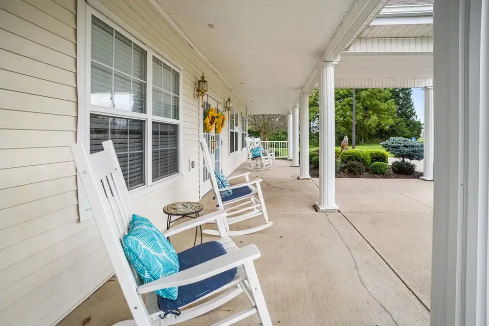 A covered porch at Trustwell Senior Living features white rocking chairs with blue cushions, a small glass table, and decorative sunflowers on the wall, all overlooking a beautifully landscaped yard.