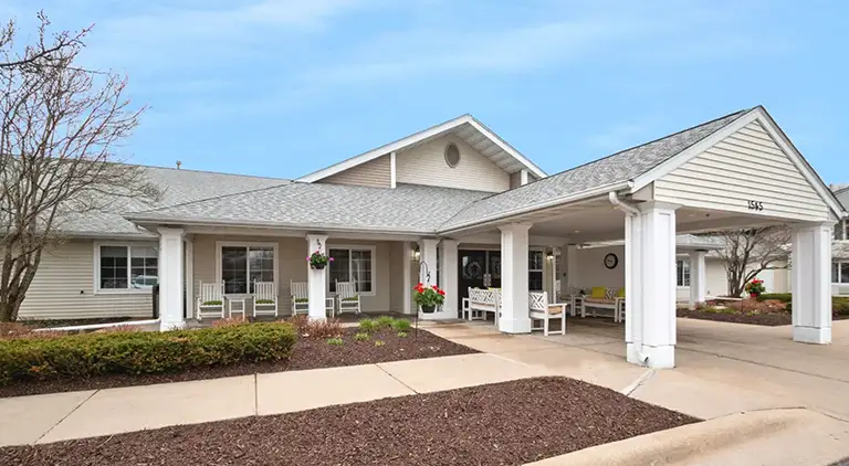 Single-story Trustwell Senior Living building with a covered entrance, white pillars, benches, flower baskets, and a landscaped walkway under a partly cloudy sky.
