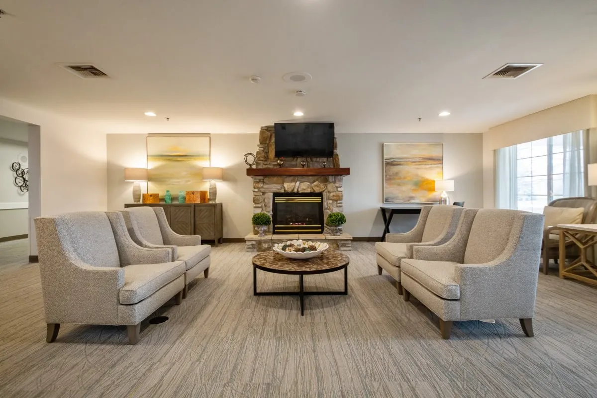 A modern lounge at Trustwell Senior Living features four beige armchairs, a round coffee table, a stone fireplace with a TV above it, and two landscape paintings on the wall.