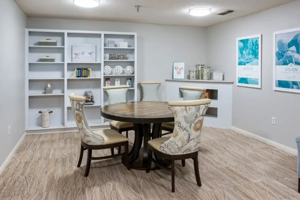 A round wooden table with four patterned chairs is set in a light-colored room at Trustwell Senior Living, featuring built-in shelves and two framed posters on the wall.