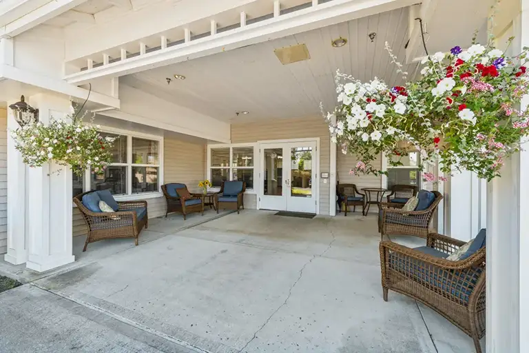 Covered porch area with wicker seating, cushions, and hanging flower baskets welcomes you at the glass doors leading to the Trustwell Senior Living entrance. Covered porch area with wicker seating, cushions, and hanging flower baskets welcomes you at the glass doors leading to the Trustwell Senior Living entrance.