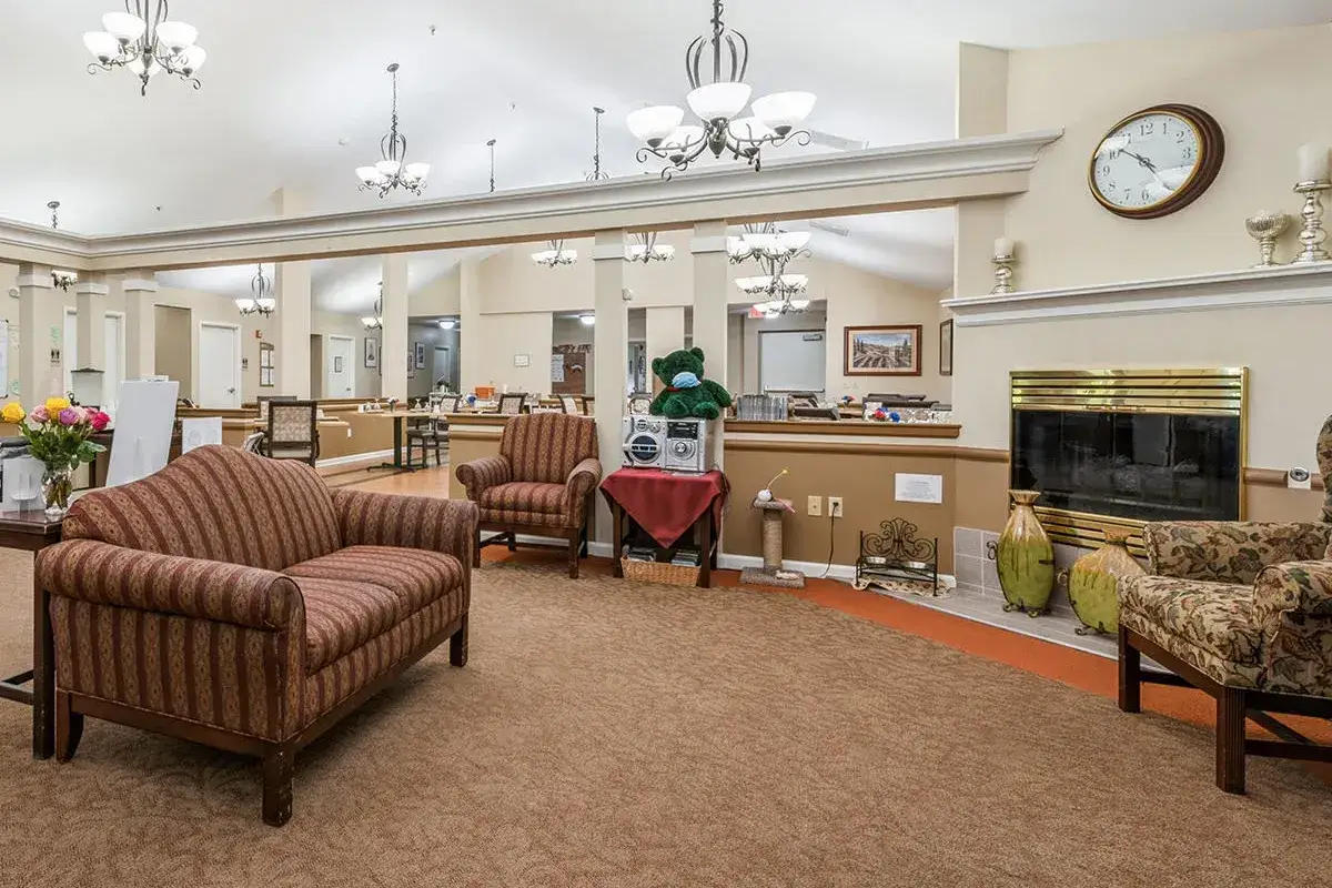 A spacious, well-lit lounge at Trustwell Senior Living features patterned armchairs, a fireplace with a clock above it, decorative vases, and mirrors along the back wall.