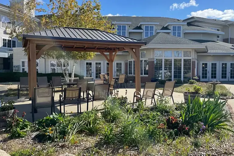 A patio with tables and chairs sits under a wooden gazebo outside a Trustwell Senior Living community, surrounded by lush plants and beautiful landscaping.