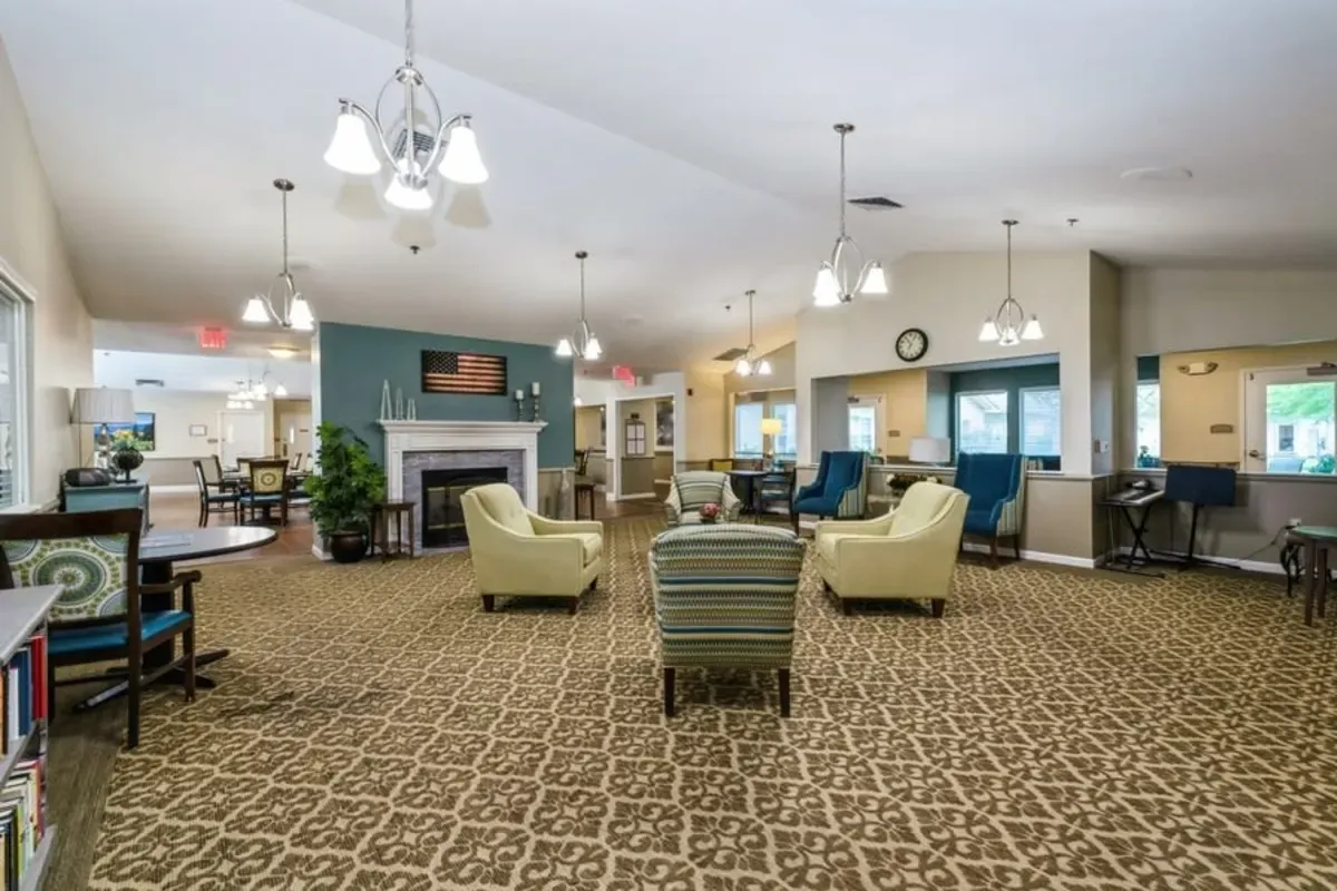 A spacious Trustwell Senior Living lounge area with patterned carpet, armchairs, desks, wall clocks, ceiling lights, and a fireplace adorned with an American flag display.