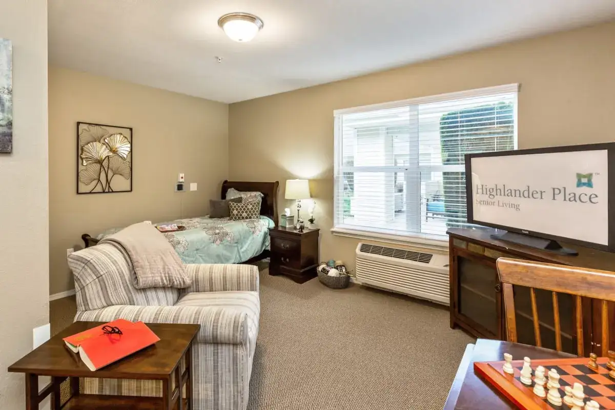 A well-lit Trustwell Senior Living apartment with a single bed, armchair, side table, TV, window, and small desk with a chessboard.