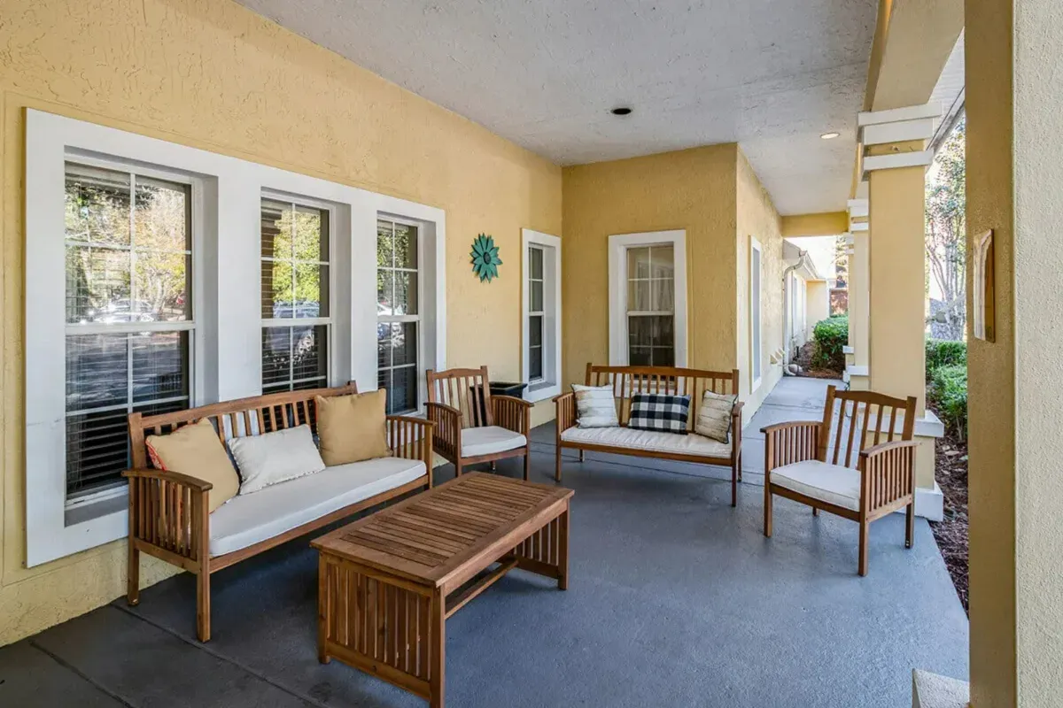 A covered patio with yellow walls at Trustwell Senior Living features two wooden loveseats, two chairs, a coffee table, and decorative cushions arranged around the seating area.