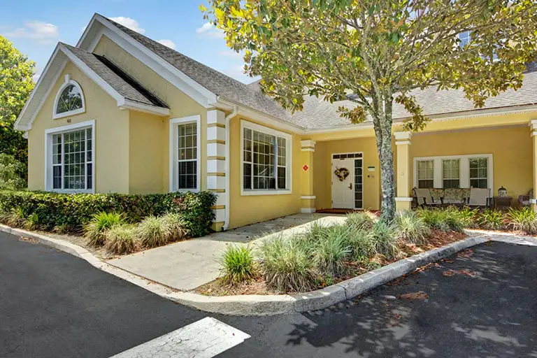 Charming single-story yellow house with white trim and a gabled roof, featuring a front porch and lush landscaping next to a paved driveway—an inviting example of Trustwell Senior Living comfort.