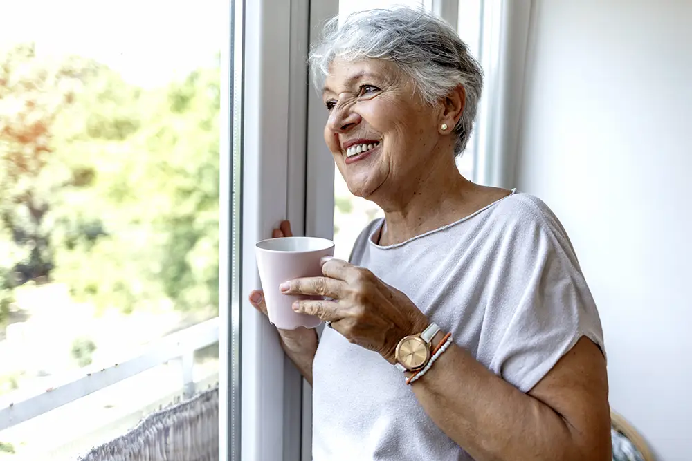 Older woman with short gray hair stands by a window holding a white mug, smiling and looking outside. She wears a light top and a watch. Greenery is visible through the window.