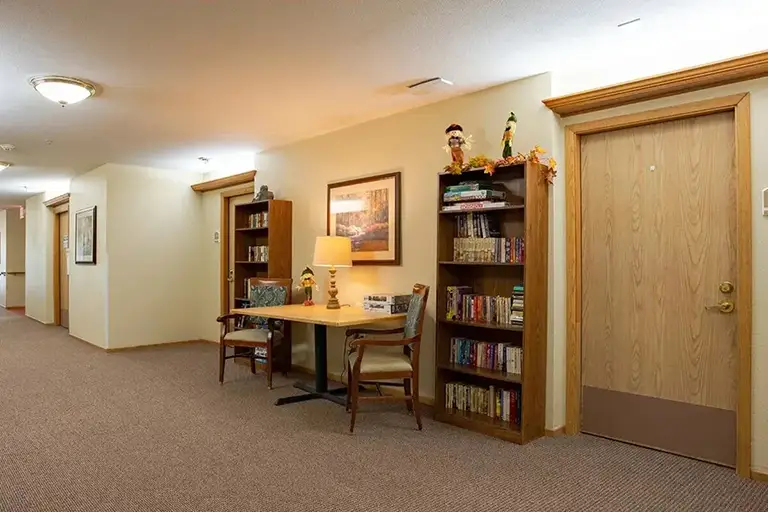 A hallway at Trustwell Senior Living features a carpeted floor, two bookshelves, a table with two chairs, a lamp, framed artwork, and a closed wooden door. A hallway at Trustwell Senior Living features a carpeted floor, two bookshelves, a table with two chairs, a lamp, framed artwork, and a closed wooden door.