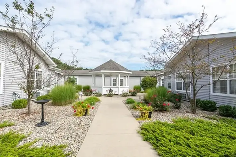 A concrete walkway leads to a white gazebo surrounded by shrubs, flowers, and two single-story gray Trustwell Senior Living buildings under a partly cloudy sky. A concrete walkway leads to a white gazebo surrounded by shrubs, flowers, and two single-story gray Trustwell Senior Living buildings under a partly cloudy sky.