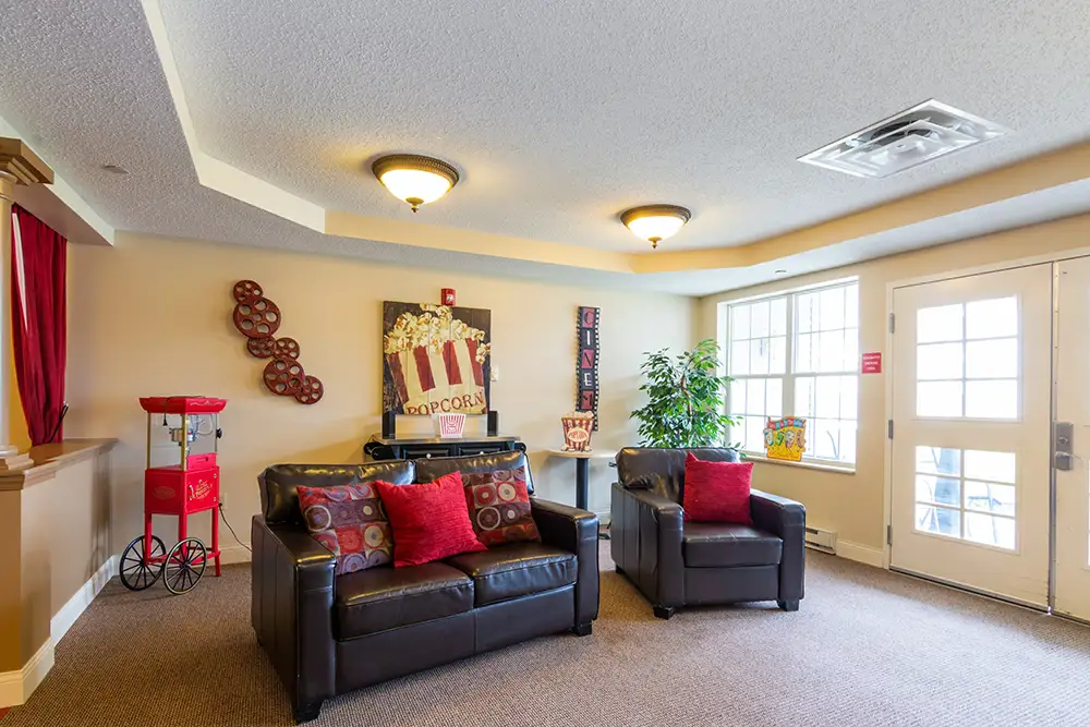 A cozy room at Trustwell Senior Living features two black armchairs, a popcorn machine, popcorn-themed decor, a large window, and a door leading outside.