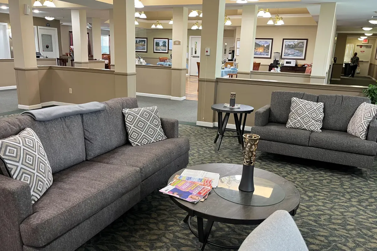 A communal lounge area at Trustwell Senior Living, featuring two gray sofas, a coffee table with magazines and a vase, patterned carpet, and framed pictures on the walls in the background.