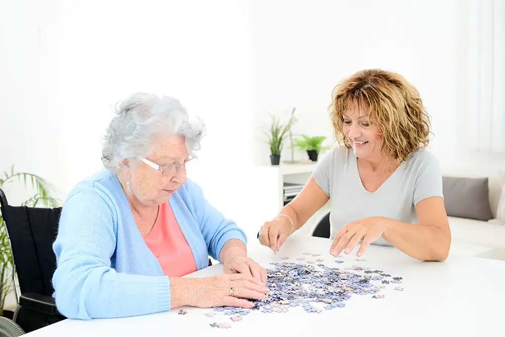 An elderly woman and a younger woman sit at a table assembling a jigsaw puzzle together in a bright room.