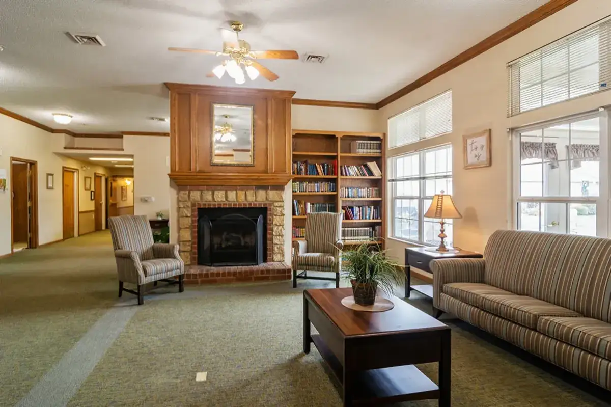 A cozy lounge area at Trustwell Senior Living features a couch, armchairs, coffee table, fireplace, bookshelf, table lamp, and large windows that fill the space with natural light.