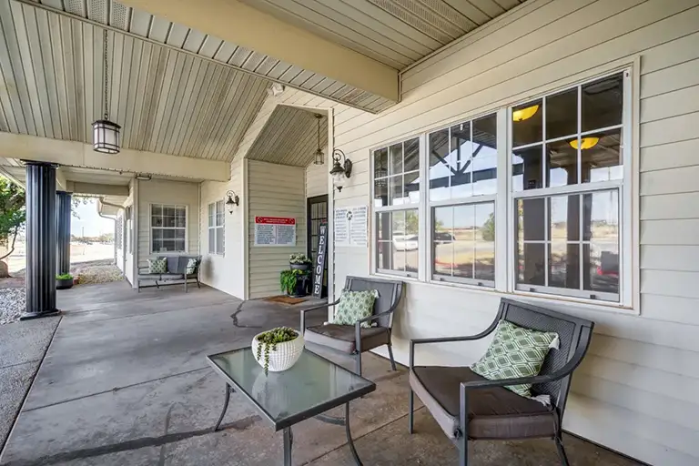 Covered outdoor seating area with two chairs, a glass table, and potted plants welcomes you in front of a Trustwell Senior Living building with large windows and signage on the wall. Covered outdoor seating area with two chairs, a glass table, and potted plants welcomes you in front of a Trustwell Senior Living building with large windows and signage on the wall.