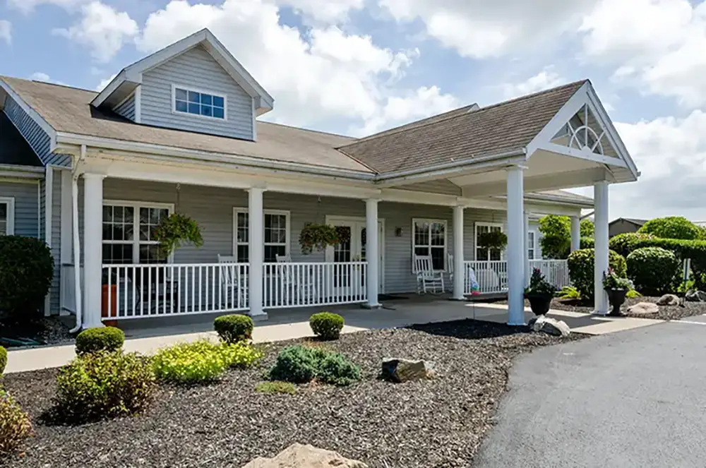Single-story Trustwell Senior Living building with a covered entrance, white pillars, front porch, shrub landscaping, and a driveway under a partly cloudy sky.