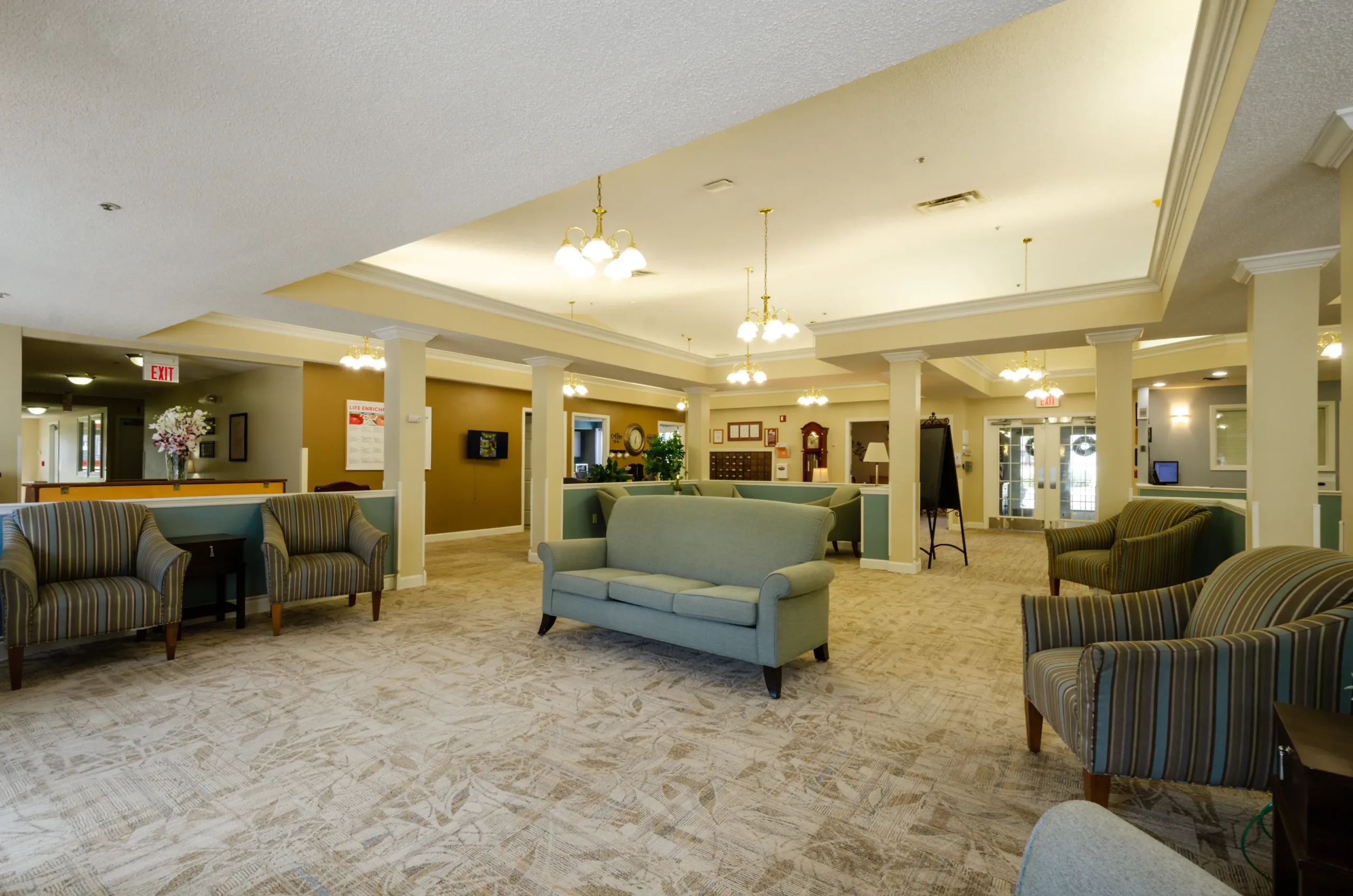 Spacious lobby area at Trustwell Senior Living, featuring a carpeted floor, green sofa, striped armchairs, decorative pillars, and ceiling lights. Reception desk and exit sign are visible in the background.