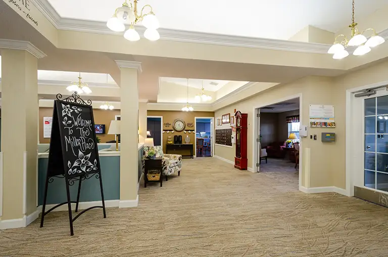 Spacious lobby area at Trustwell Senior Living with beige walls, carpeted floor, overhead lights, a welcome sign on an easel, comfortable seating, and decorative elements. Multiple doorways lead to other rooms. Spacious lobby area at Trustwell Senior Living with beige walls, carpeted floor, overhead lights, a welcome sign on an easel, comfortable seating, and decorative elements. Multiple doorways lead to other rooms.