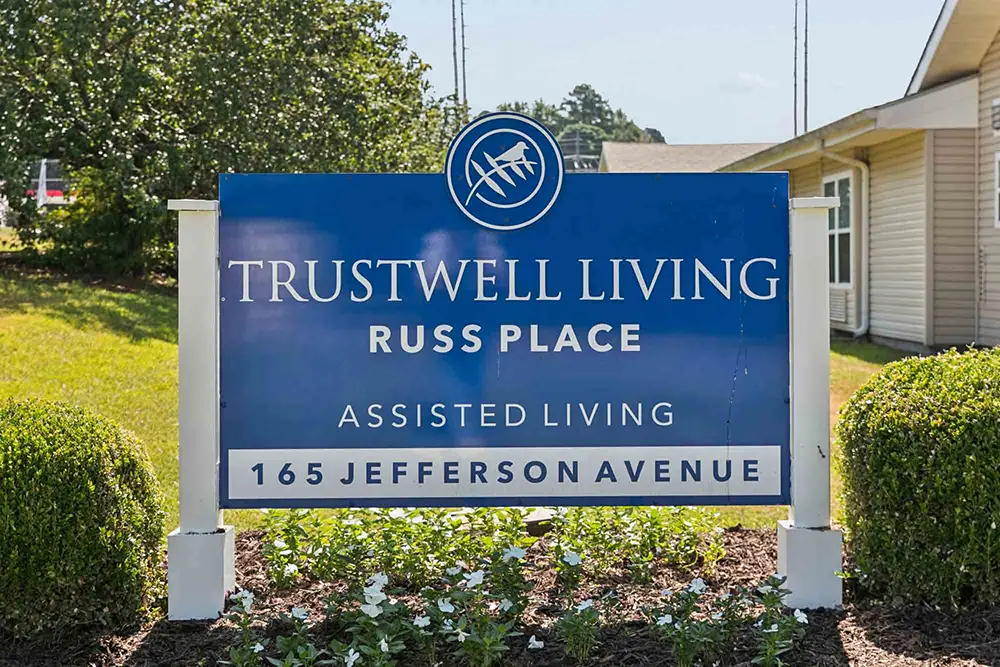 Blue and white sign reads “Trustwell Living Russ Place Assisted Living, 165 Jefferson Avenue” in front of a building with greenery and flowers. Blue and white sign reads “Trustwell Living Russ Place Assisted Living, 165 Jefferson Avenue” in front of a building with greenery and flowers.