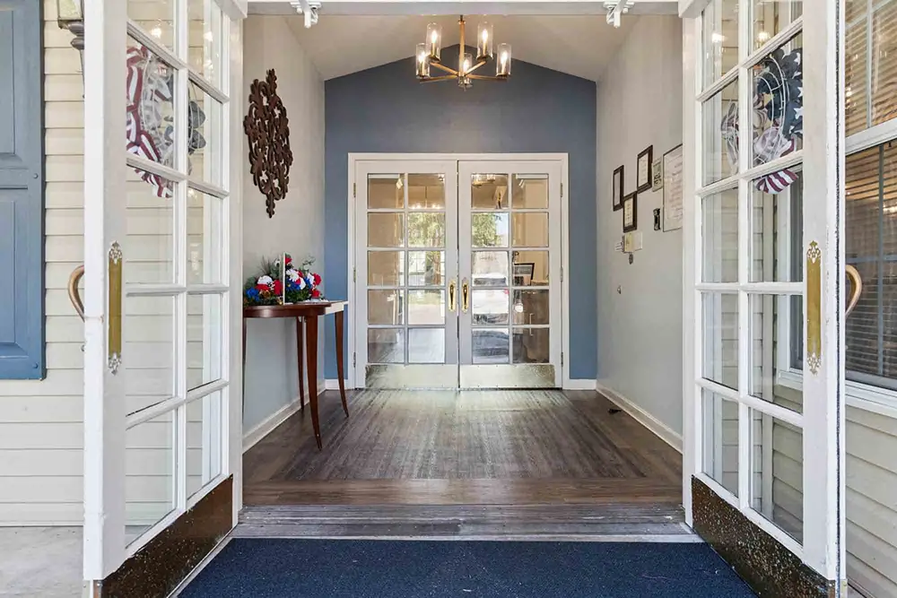 Double glass doors lead to a hallway with wood floors, a side table with flowers, wall art, a chandelier, and more glass doors at the end. Double glass doors lead to a hallway with wood floors, a side table with flowers, wall art, a chandelier, and more glass doors at the end.