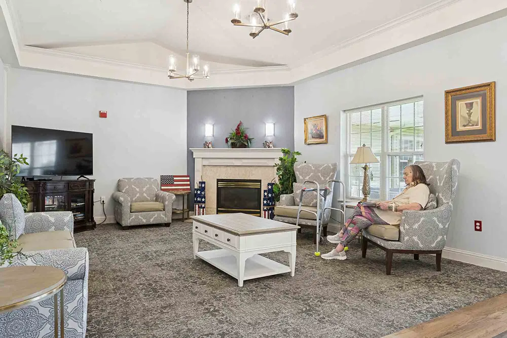 A woman sits reading in a bright, neatly furnished living room with armchairs, a fireplace, a TV, and patriotic decor. A woman sits reading in a bright, neatly furnished living room with armchairs, a fireplace, a TV, and patriotic decor.