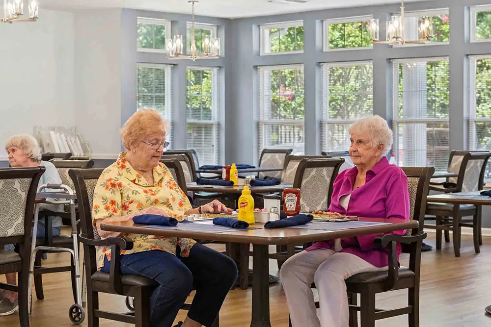 Two elderly women sit at a dining table in a bright, spacious room with large windows, condiments, and napkins on the table. Other people are seated in the background. Two elderly women sit at a dining table in a bright, spacious room with large windows, condiments, and napkins on the table. Other people are seated in the background.