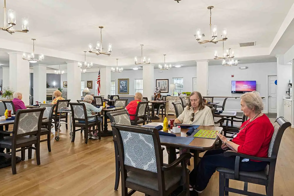 Several elderly people sit at tables in a bright, spacious common room with chandeliers, playing games and socializing. An American flag is visible in the background. Several elderly people sit at tables in a bright, spacious common room with chandeliers, playing games and socializing. An American flag is visible in the background.