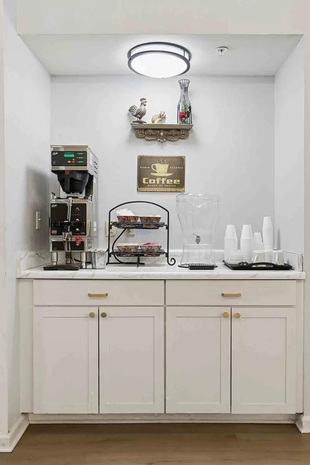 Coffee station with a coffee maker, cups, water dispenser, and a tiered tray of condiments on a white counter beneath a shelf with decorative items and a “Coffee” sign. Coffee station with a coffee maker, cups, water dispenser, and a tiered tray of condiments on a white counter beneath a shelf with decorative items and a “Coffee” sign.