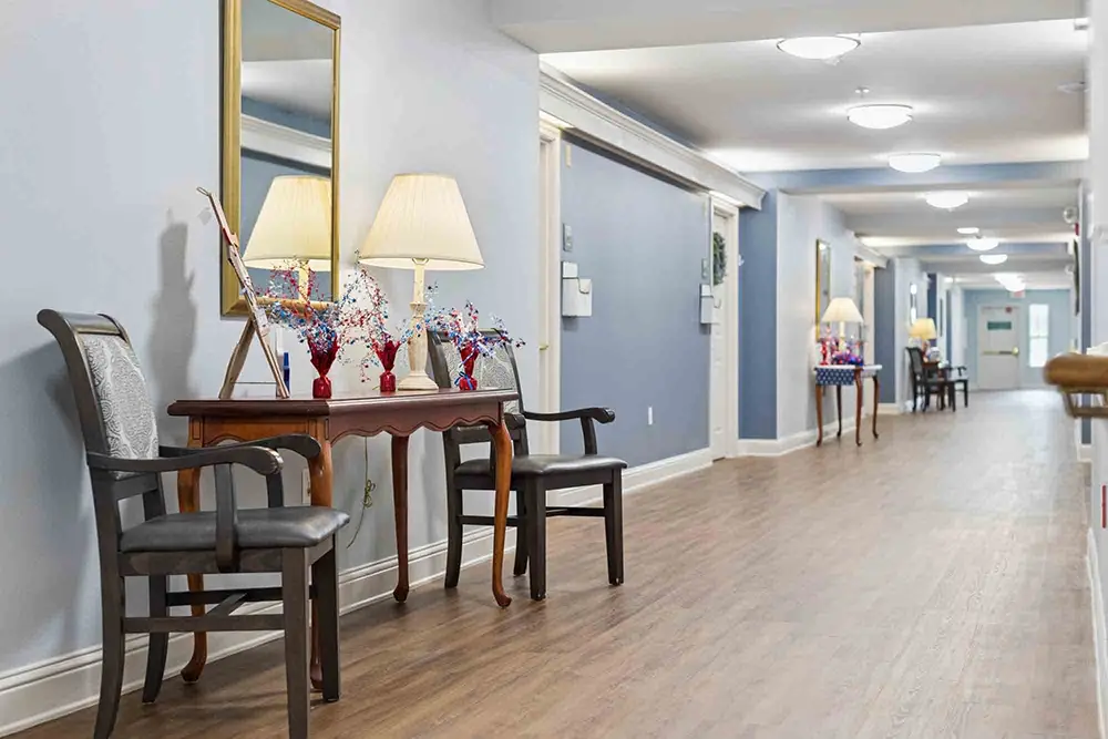 A hallway with wood flooring, decorative tables, chairs, lamps, mirrors, and patriotic centerpieces along a light blue wall. A hallway with wood flooring, decorative tables, chairs, lamps, mirrors, and patriotic centerpieces along a light blue wall.
