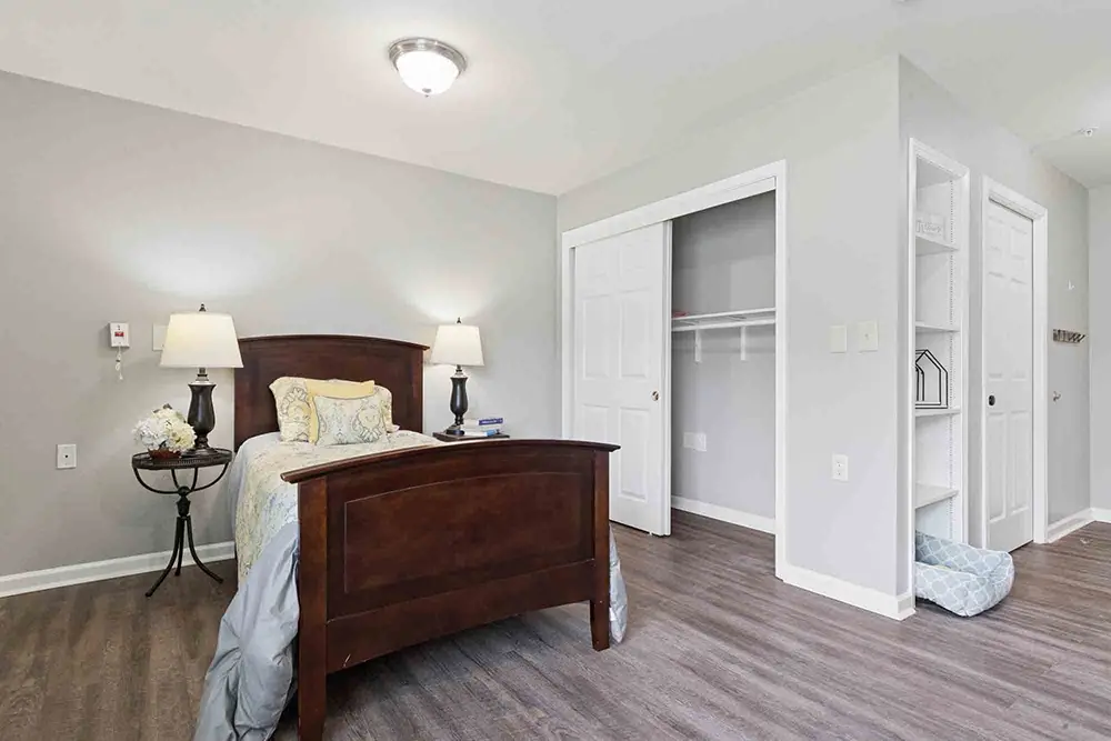 Bedroom with dark wooden bed, nightstand, two lamps, open closet with shelves, and dog bed on wood floor. Bedroom with dark wooden bed, nightstand, two lamps, open closet with shelves, and dog bed on wood floor.