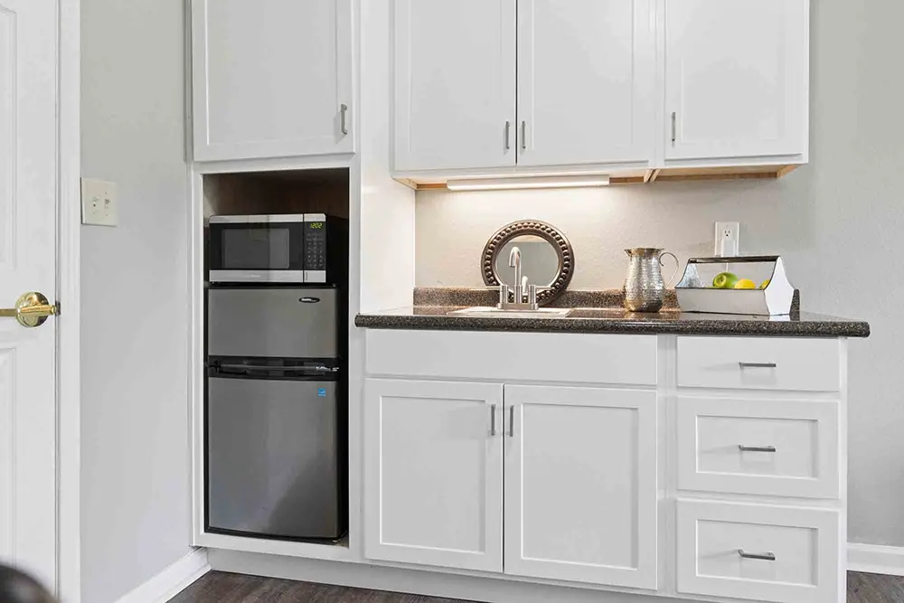 Small kitchenette with white cabinets, a countertop, a stainless steel mini fridge, a microwave, a sink, a pitcher, and a tray with lemons and apples. Small kitchenette with white cabinets, a countertop, a stainless steel mini fridge, a microwave, a sink, a pitcher, and a tray with lemons and apples.