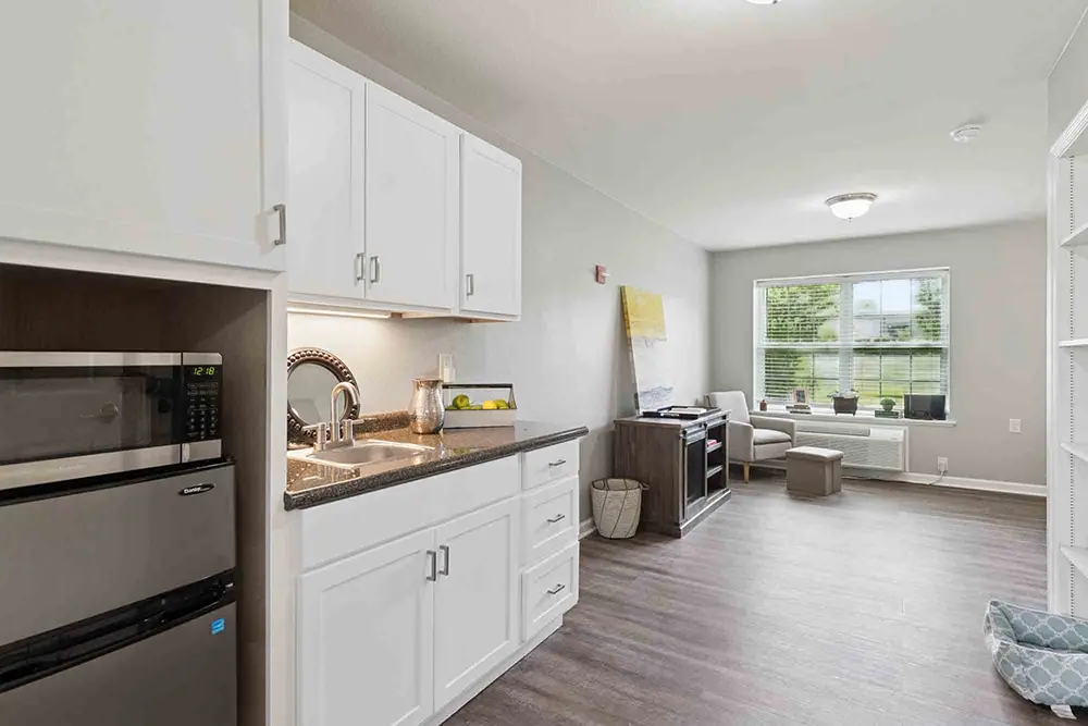 Modern studio apartment with a small kitchenette, stainless steel appliances, white cabinets, wood-style flooring, and a living area with a window and sofa in the background. Modern studio apartment with a small kitchenette, stainless steel appliances, white cabinets, wood-style flooring, and a living area with a window and sofa in the background.