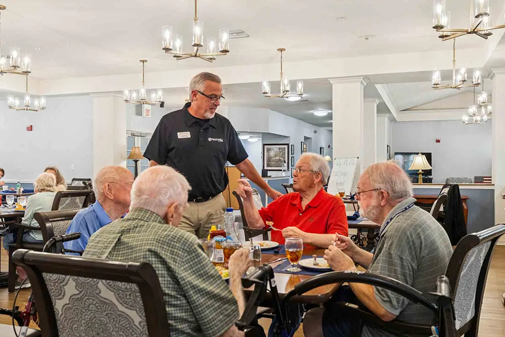 A staff member speaks with a group of older adults seated around a dining table in a well-lit communal dining area. A staff member speaks with a group of older adults seated around a dining table in a well-lit communal dining area.