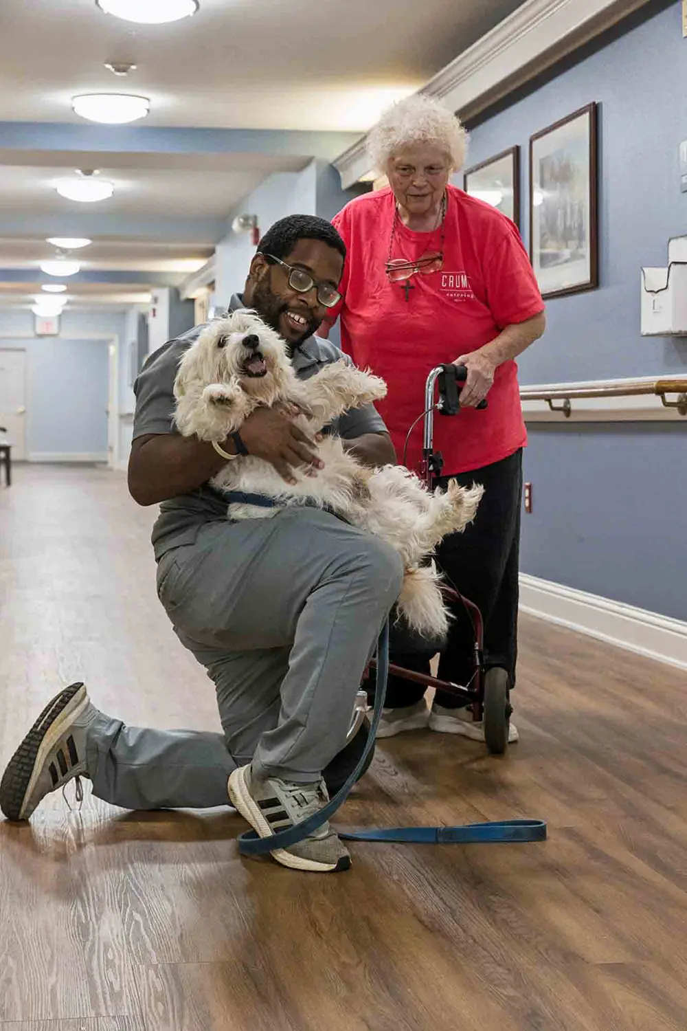 A man kneels in a hallway holding a small white dog while an older woman stands nearby with a walker, both smiling. A man kneels in a hallway holding a small white dog while an older woman stands nearby with a walker, both smiling.
