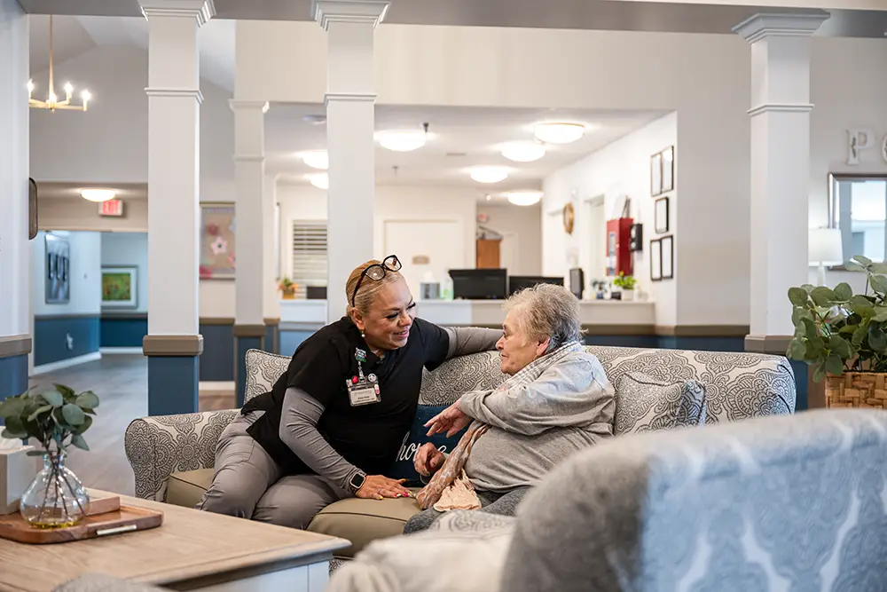 Plum Creek Place-POI-02 A healthcare worker in uniform sits on a couch, talking and smiling with an elderly woman in a care facility’s common area.