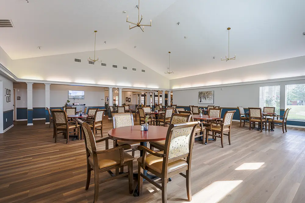 Plum Creek Place-POI-03 Spacious dining area with multiple wooden tables and chairs, large windows on one side, chandeliers hanging from a high ceiling, and bright natural lighting.