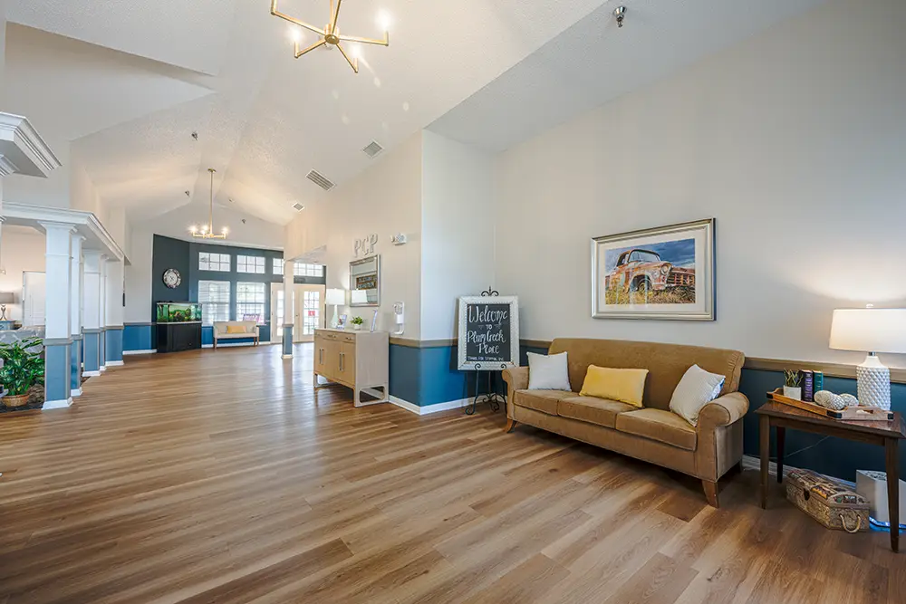 Plum Creek Place-POI-05 Spacious, well-lit lobby with wood flooring, a tan sofa, lamp, side table, wall art, and a chalkboard sign reading "Welcome" near large windows and a reception desk.