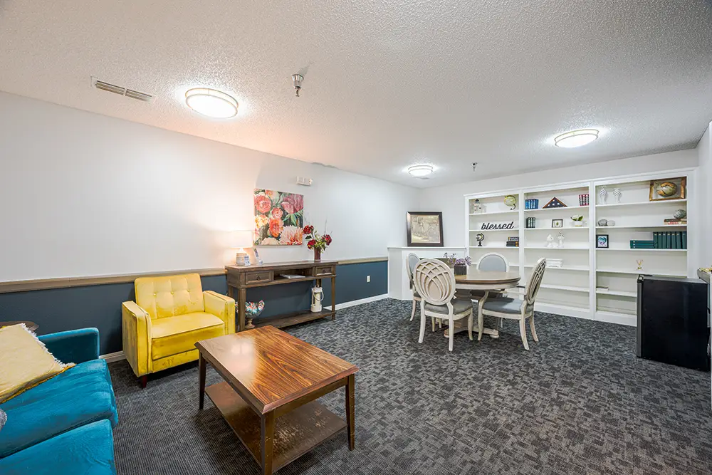 Plum Creek Place-POI-06 A brightly lit room with a yellow chair, blue sofa, wooden coffee table, white dining set, built-in shelves, and decorative artwork on the walls.